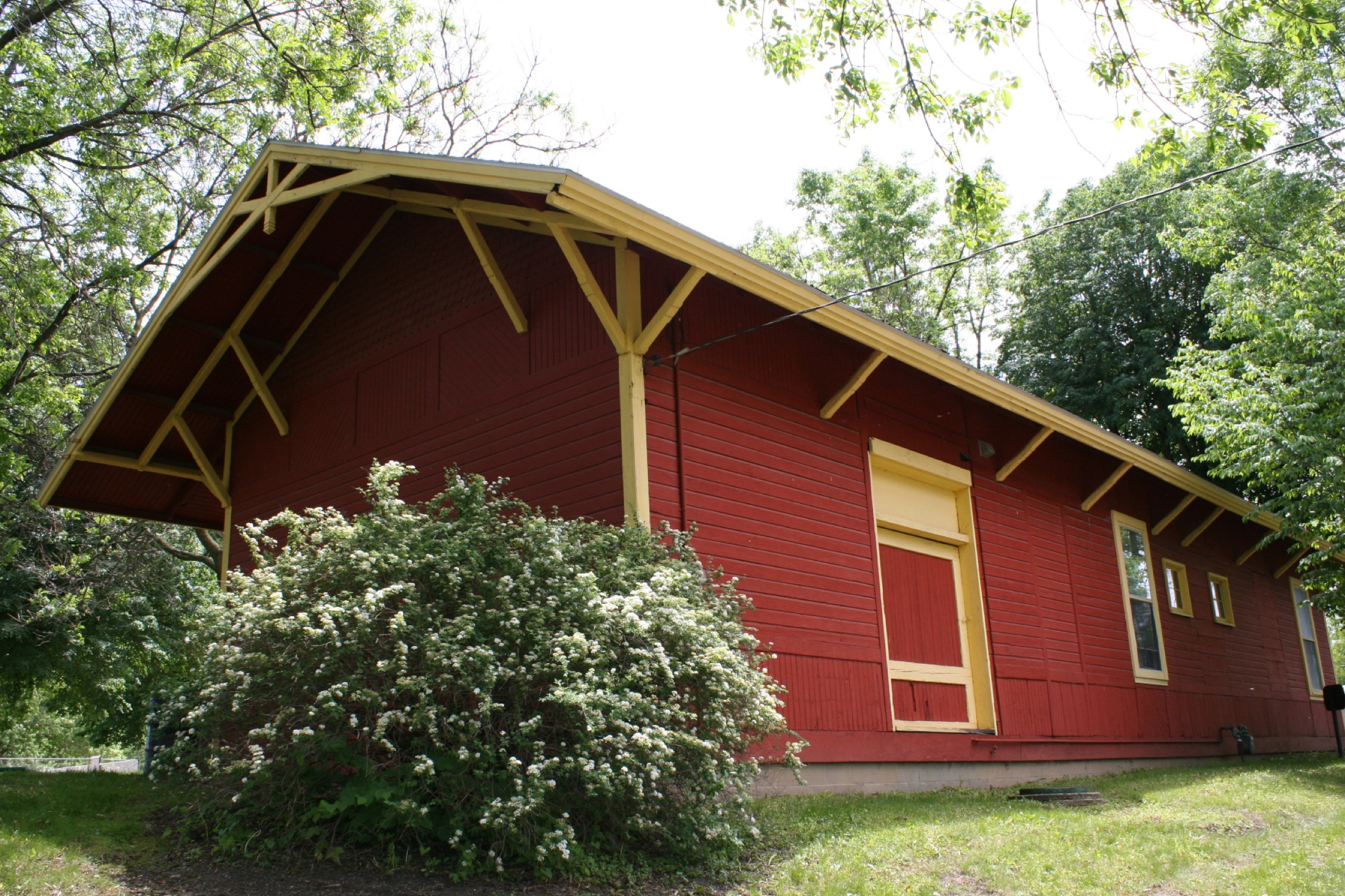 Long side of the red depot under the trees with the double-globe street lamp and a green picnic table